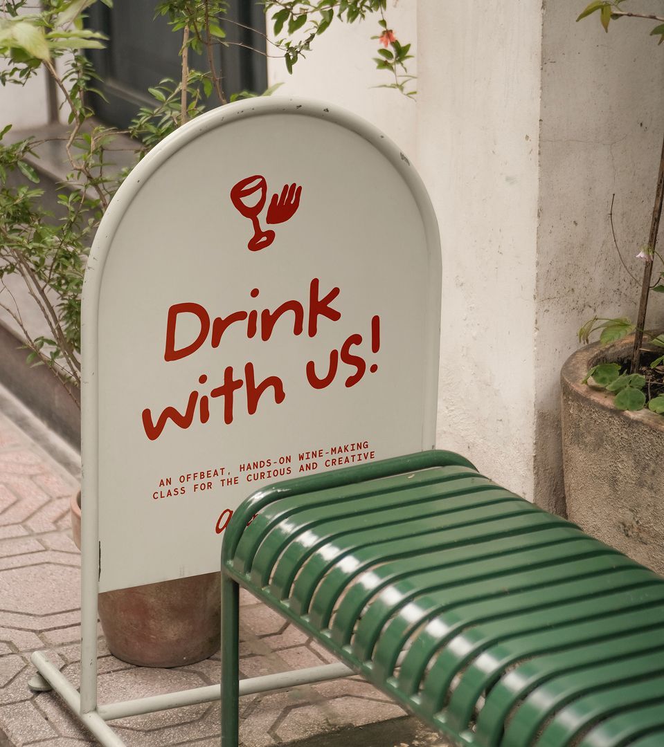 White sign with red “Drink with us!” text beside a green slatted bench outdoors