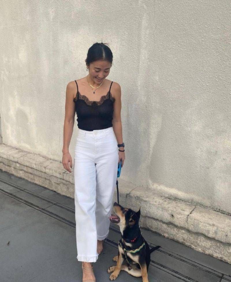 Une femme en haut noir et pantalon blanc sourit en promenant son chien en laisse.