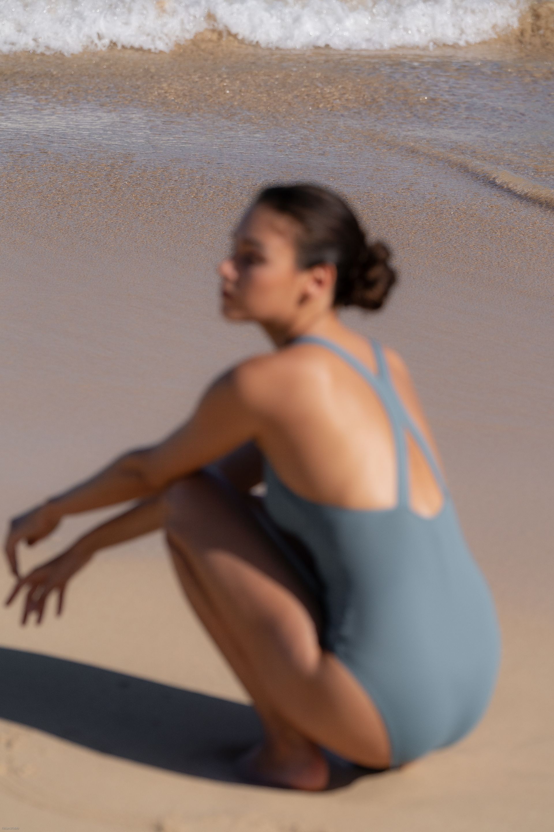 Une femme en maillot de bain bleu est accroupie sur une plage de sable fin, près de douces vagues.