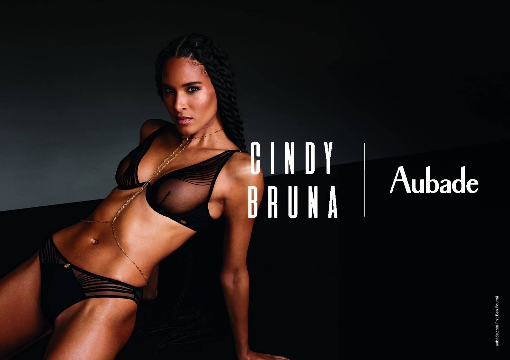 Cindy Bruna pour la lingerie Aubade. Femme en soutien-gorge et culotte noirs, chaîne de corps dorée, fond sombre.
