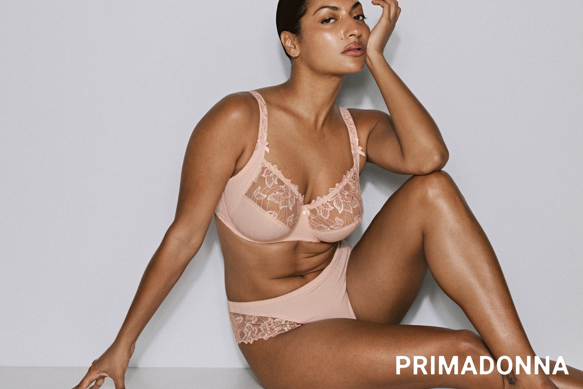 Une femme en lingerie pêche est assise, la tête inclinée, sur un fond clair.