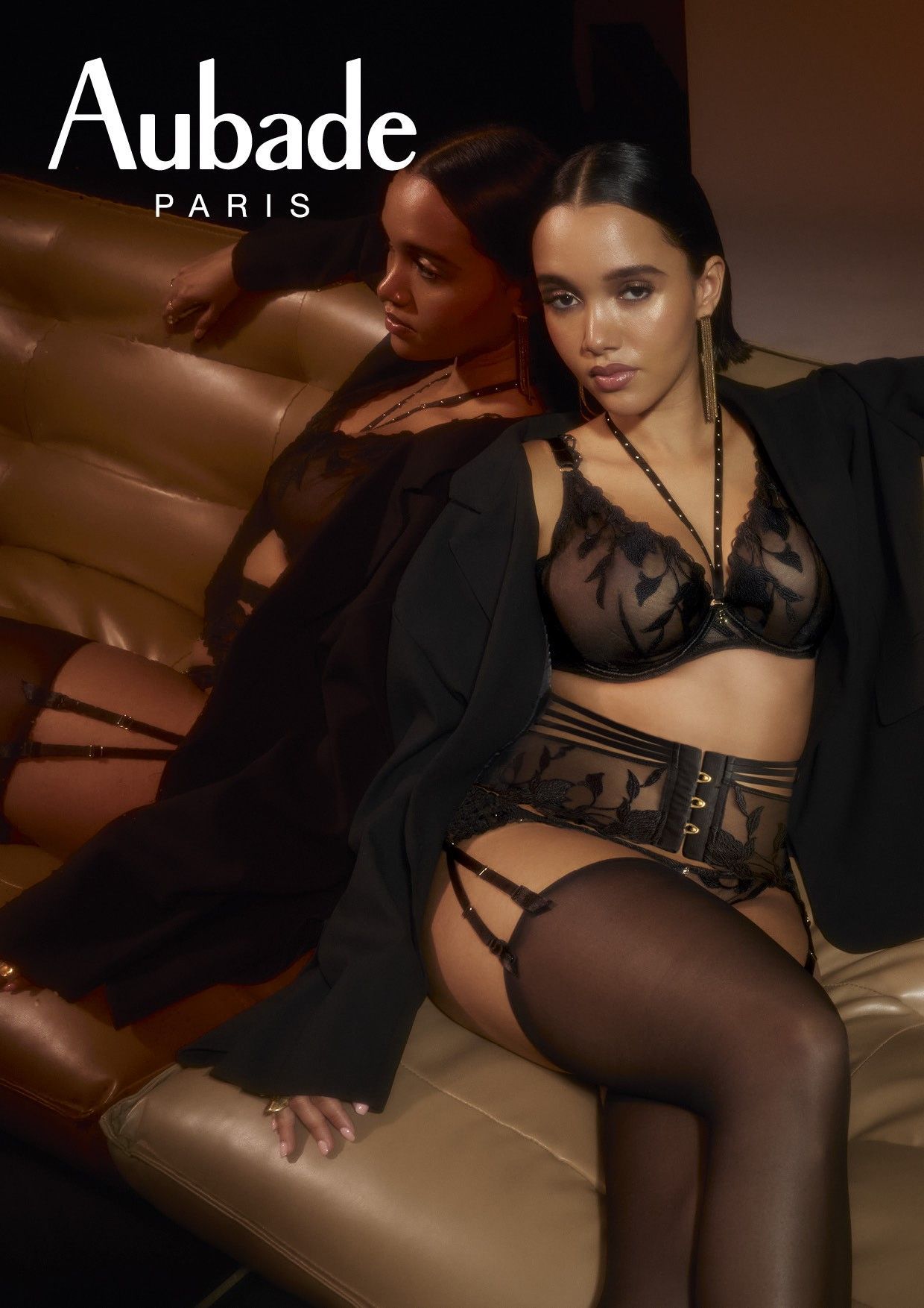 Femme en lingerie noire, blazer et bas ; assise sur un canapé beige, prenant la pose.