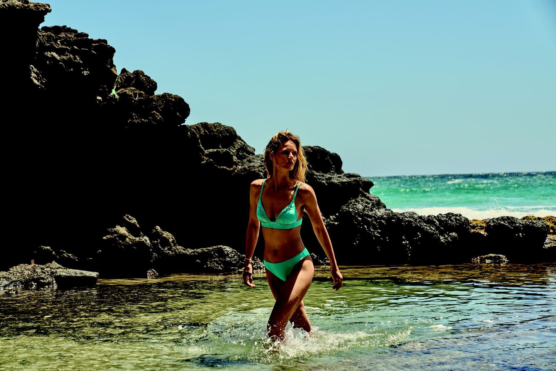 Femme en bikini turquoise pataugeant dans l'eau peu profonde près des rochers, en bord de mer.