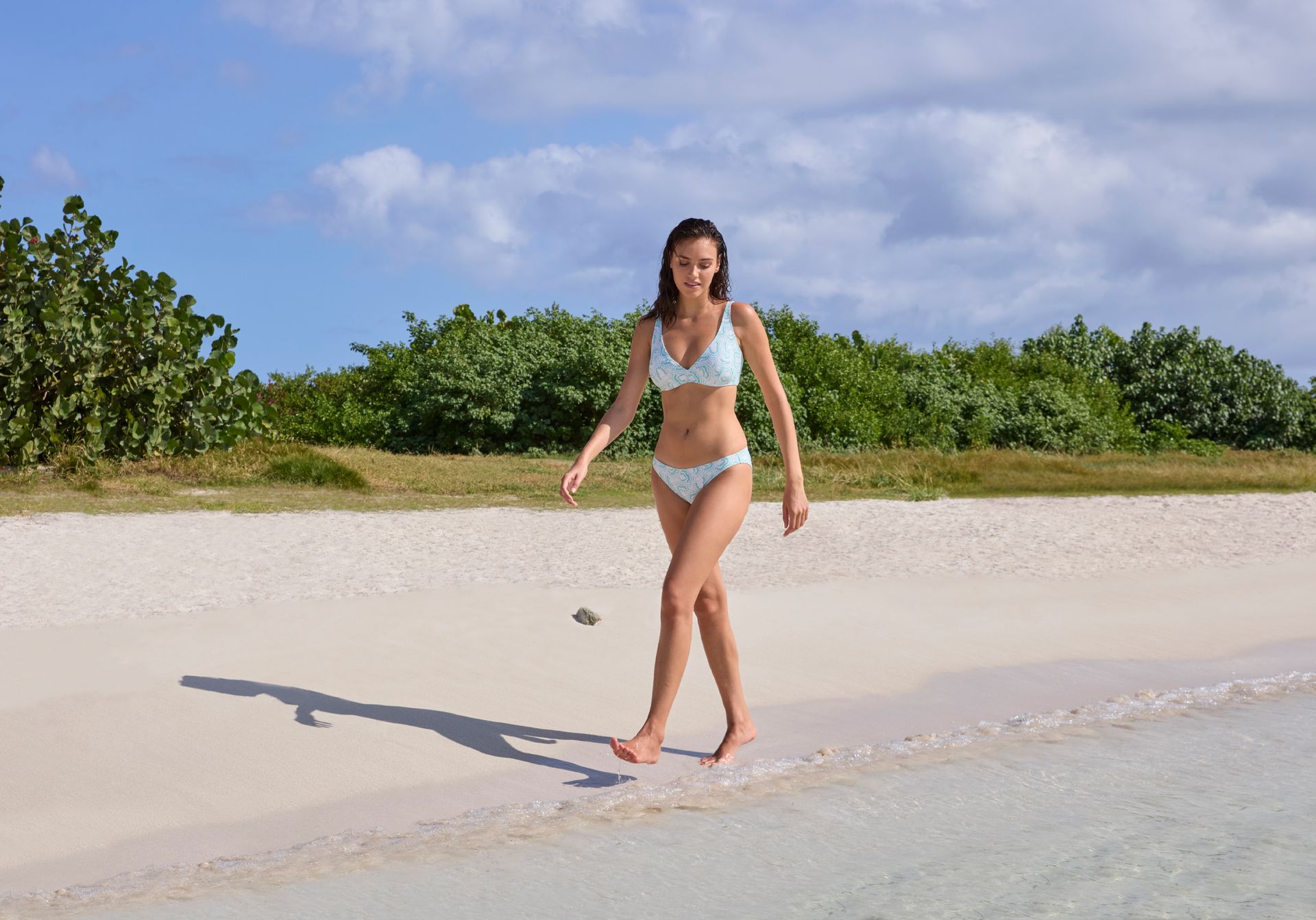 Femme en bikini marchant sur une plage de sable fin près de l'eau par une journée ensoleillée.