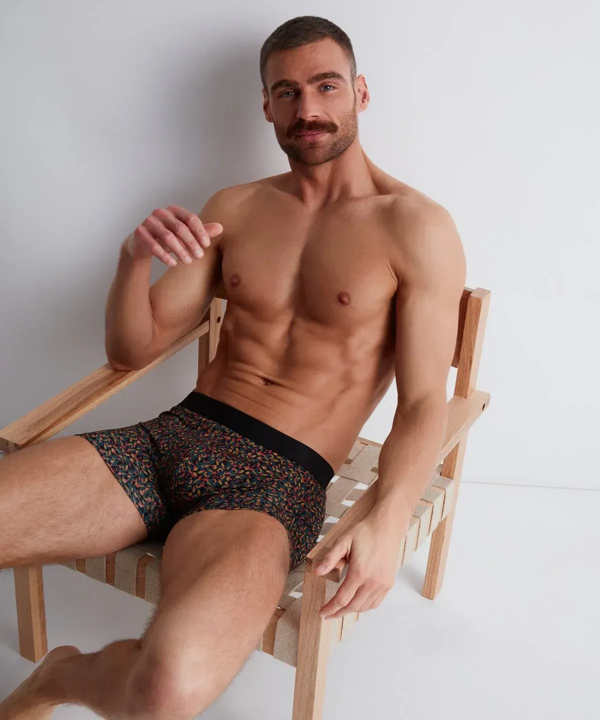 Un homme en sous-vêtements à motifs est assis, torse nu, sur une chaise en bois, souriant.