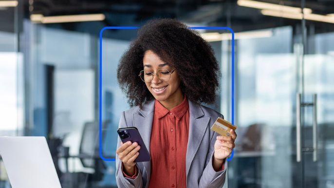A smiling Black woman in a business suit holds a phone and credit card in an office.