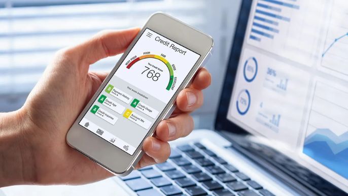 Hand holding a phone displaying a credit report with a score of 768. Laptop with charts in background.