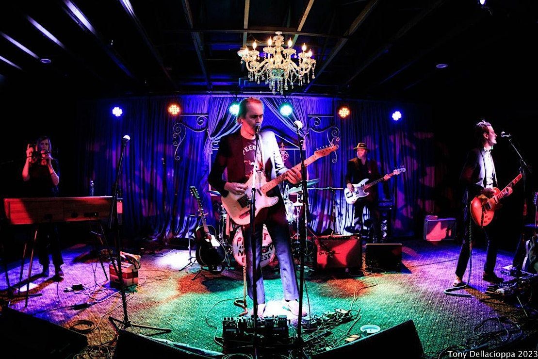 A band performs on a stage bathed in colorful lights. Musicians play guitars, keyboards, and drums in a dimly lit venue.