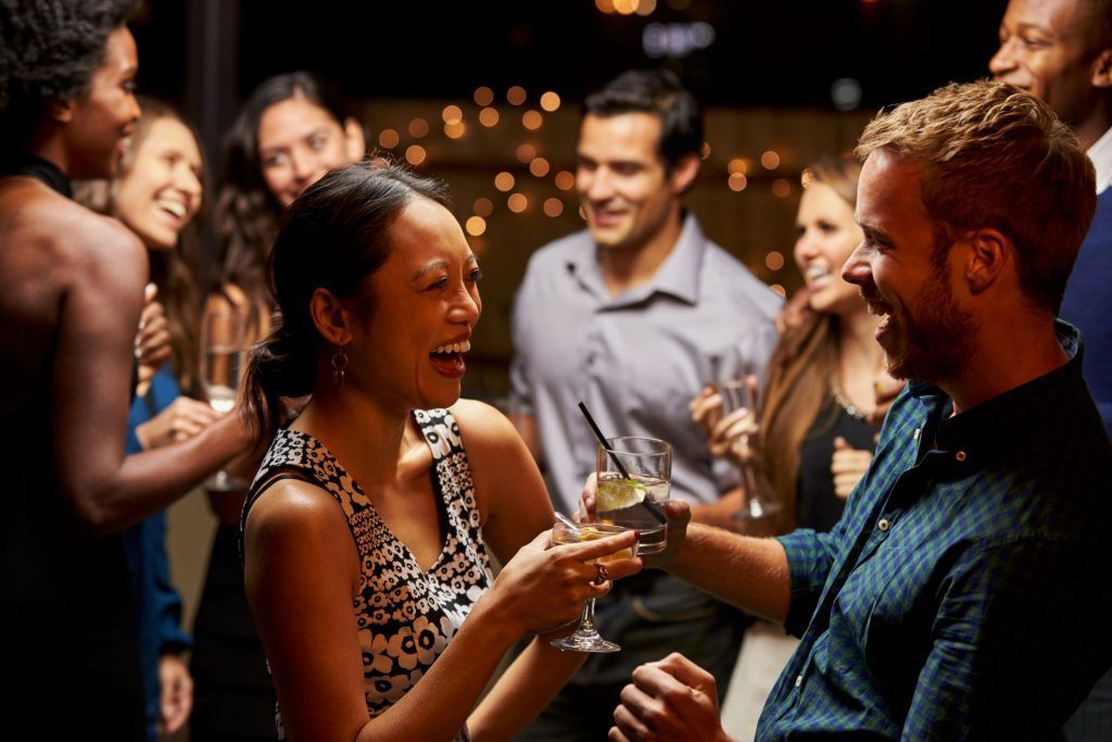 People at a party: a woman in a patterned dress laughs while offering a drink to a smiling man.