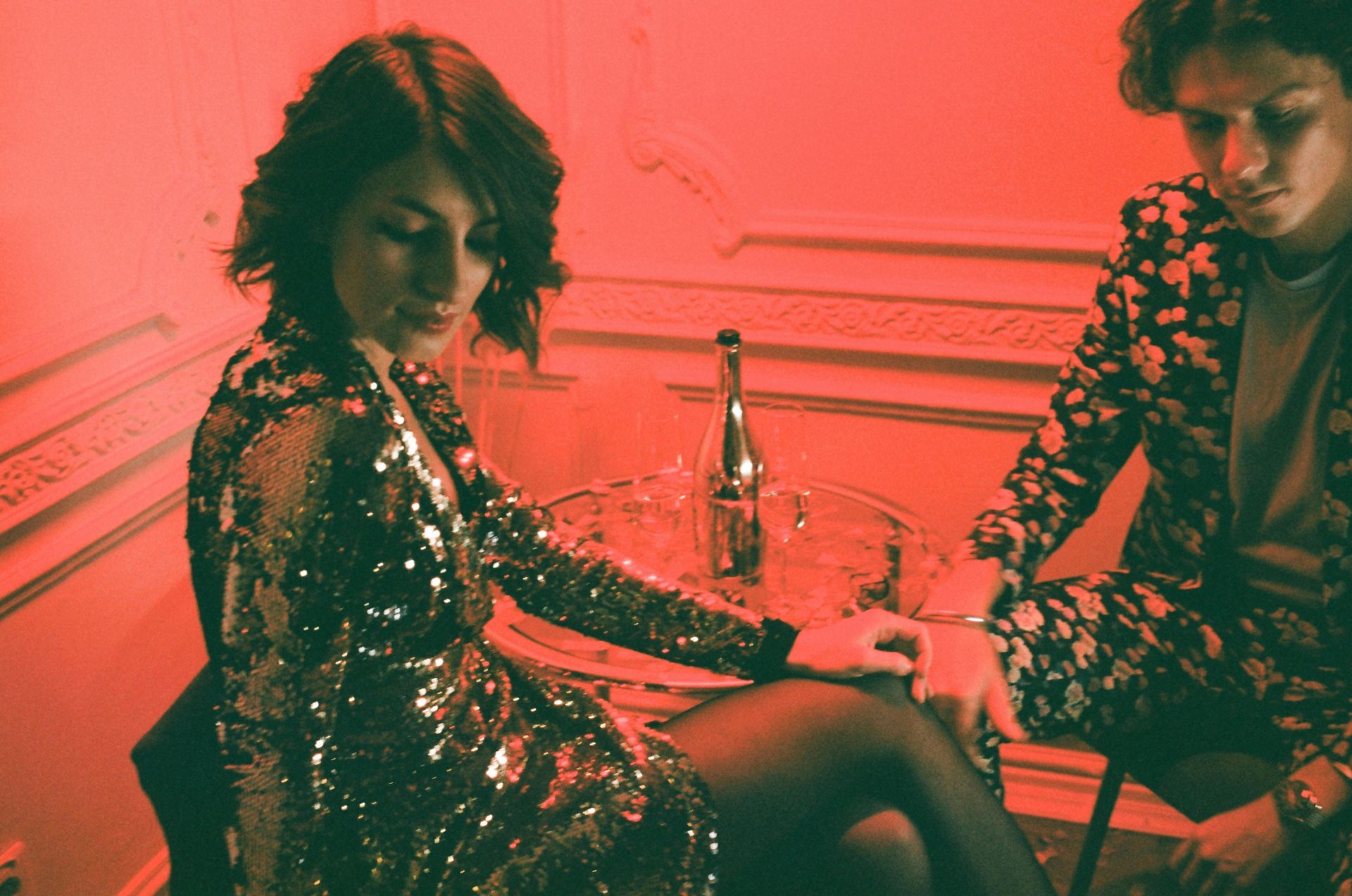 Woman in a sparkly dress and man in floral suit seated, sharing a drink at a small table, in a red-lit room.