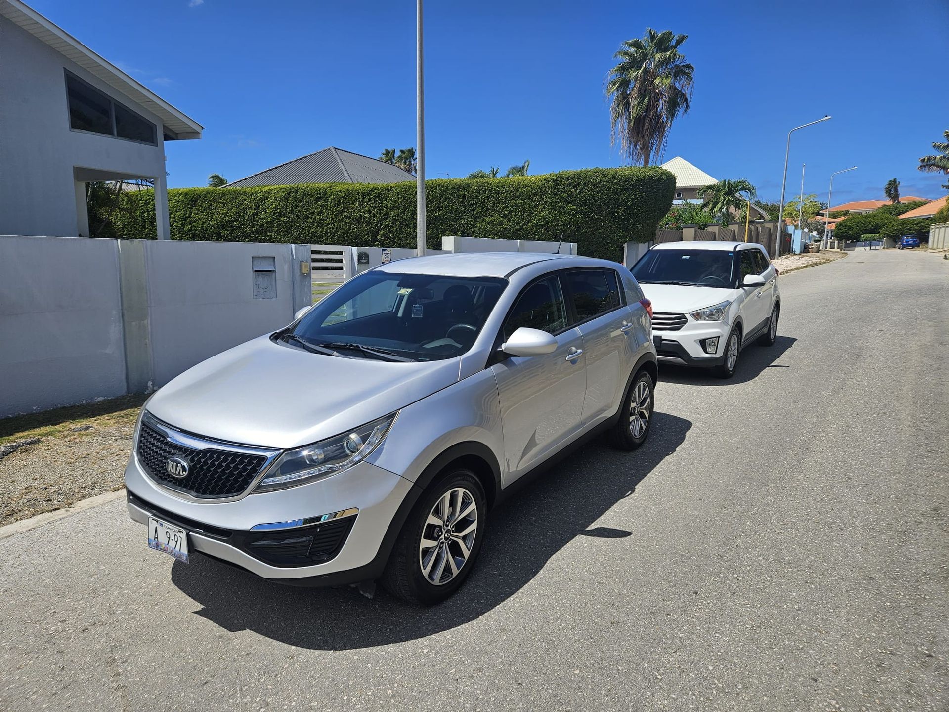 Een zilverkleurige Kia Sportage staat geparkeerd in een woonstraat achter een witte SUV onder een helderblauwe hemel.