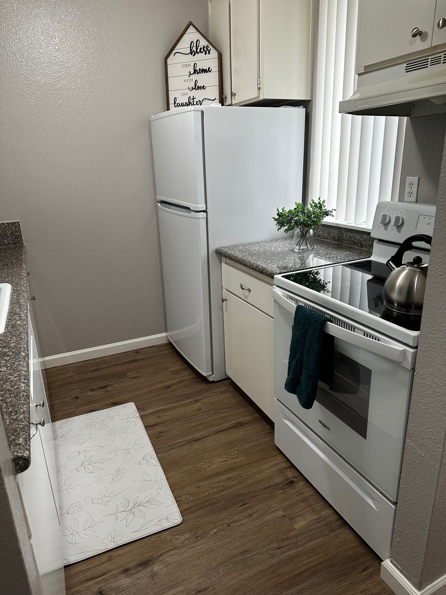 Narrow kitchen with white appliances, gray walls, wood floor, and countertop by a window.