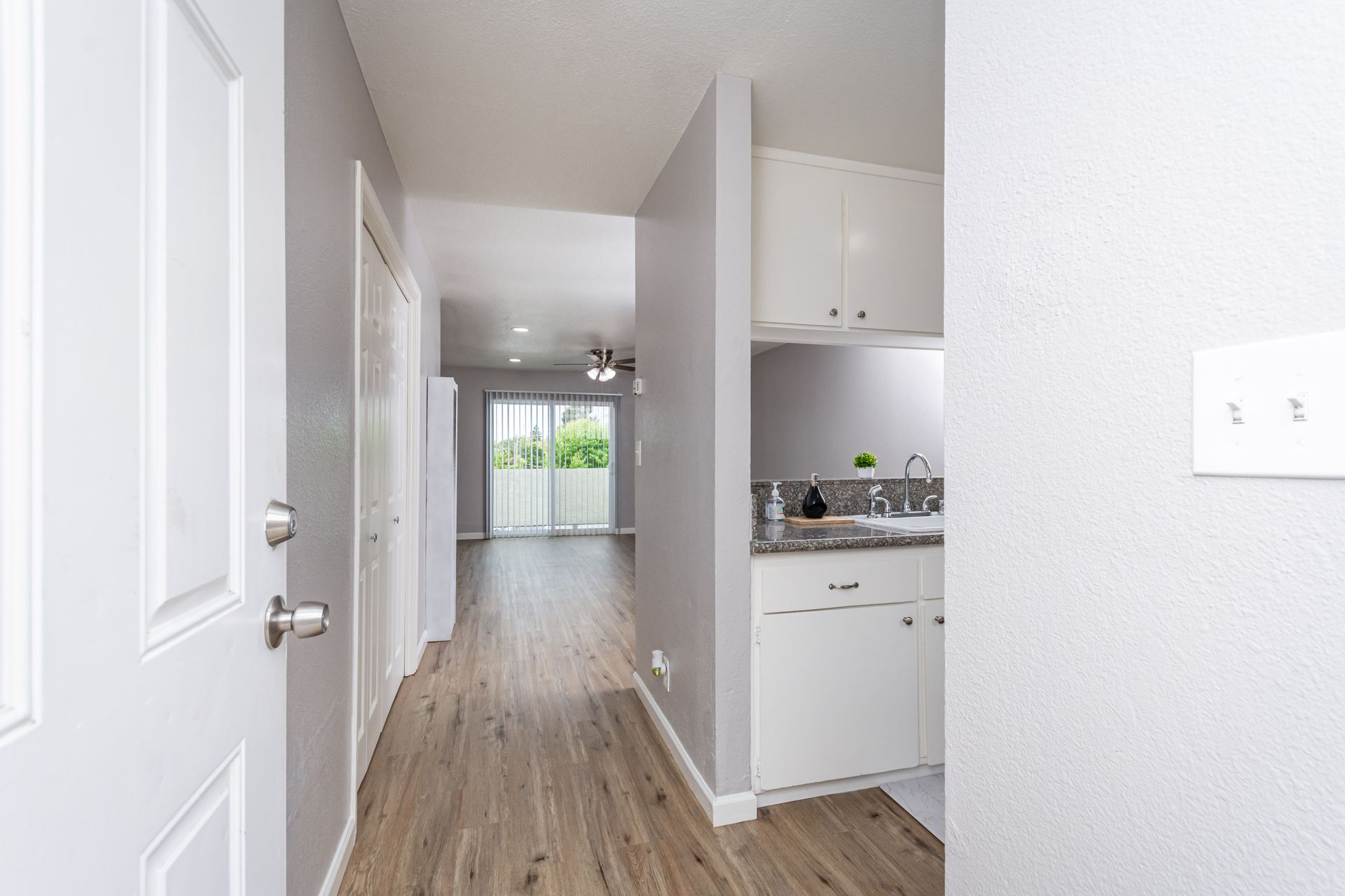 Narrow apartment hallway with white walls, wood floors, and a bright kitchen area in the distance