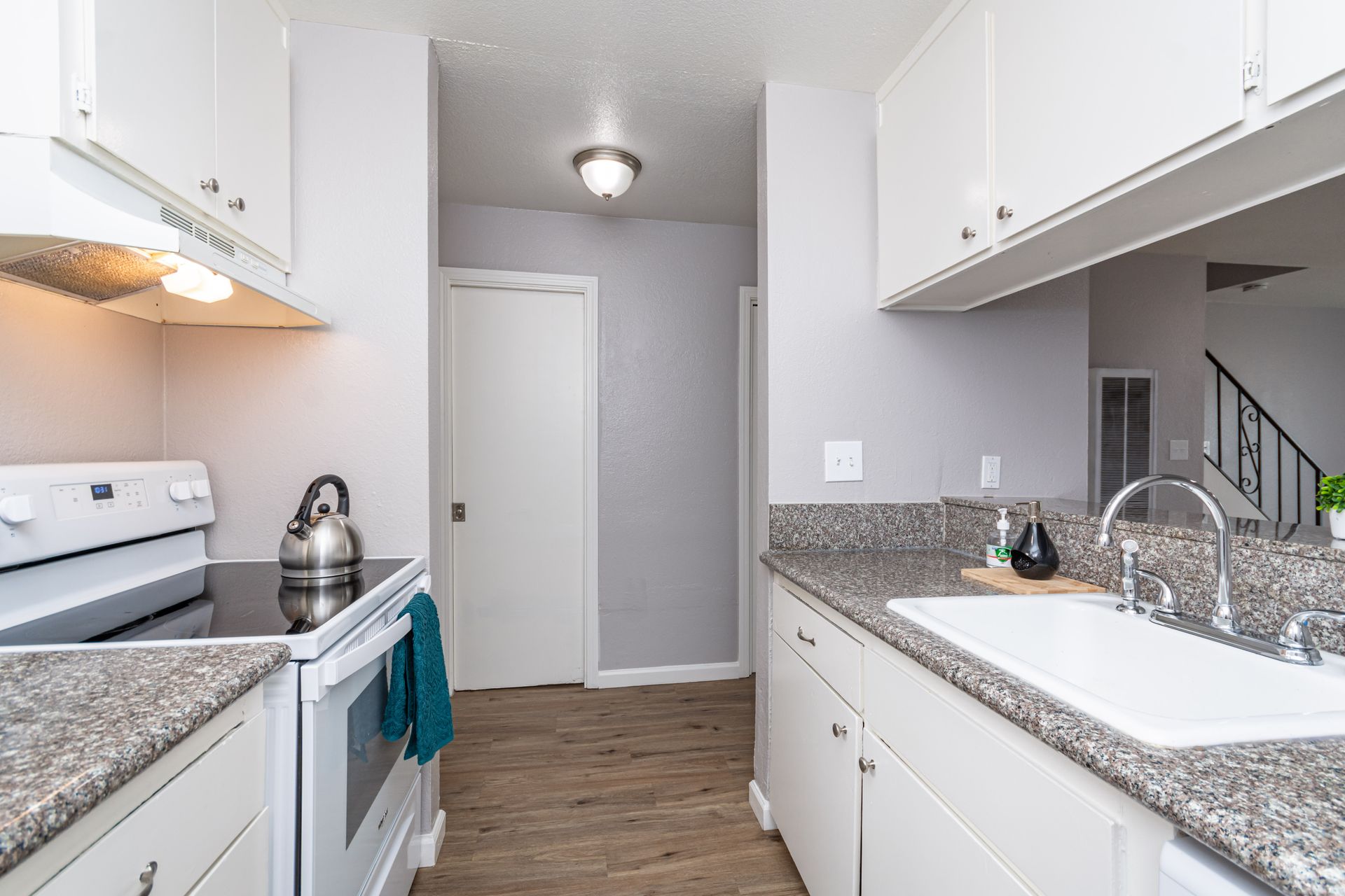 Bright white galley kitchen with granite counters, stainless stove, and sink leading to a hall doorway.