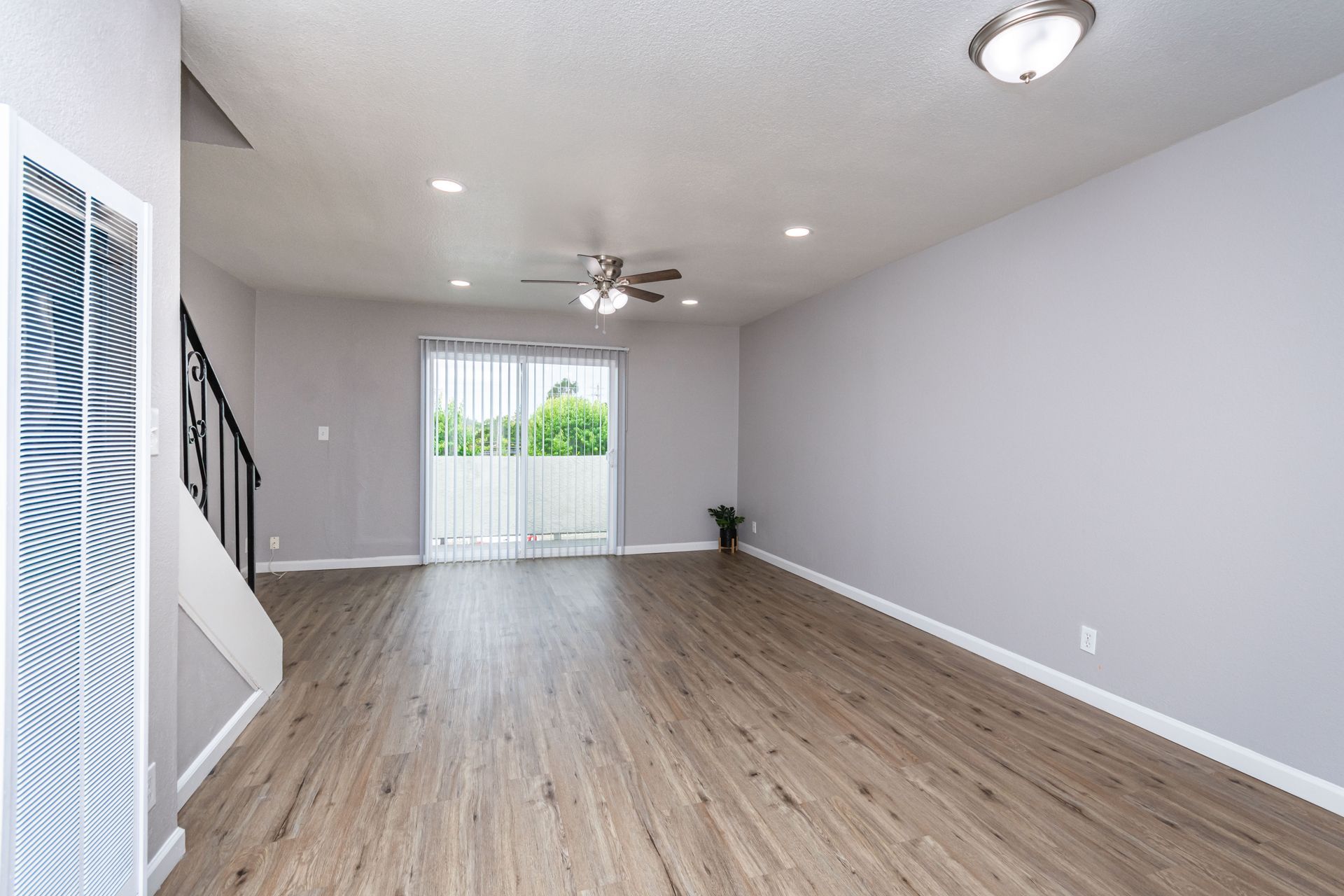 Empty modern living room with gray walls, wood floors, ceiling fan, and sliding glass doors