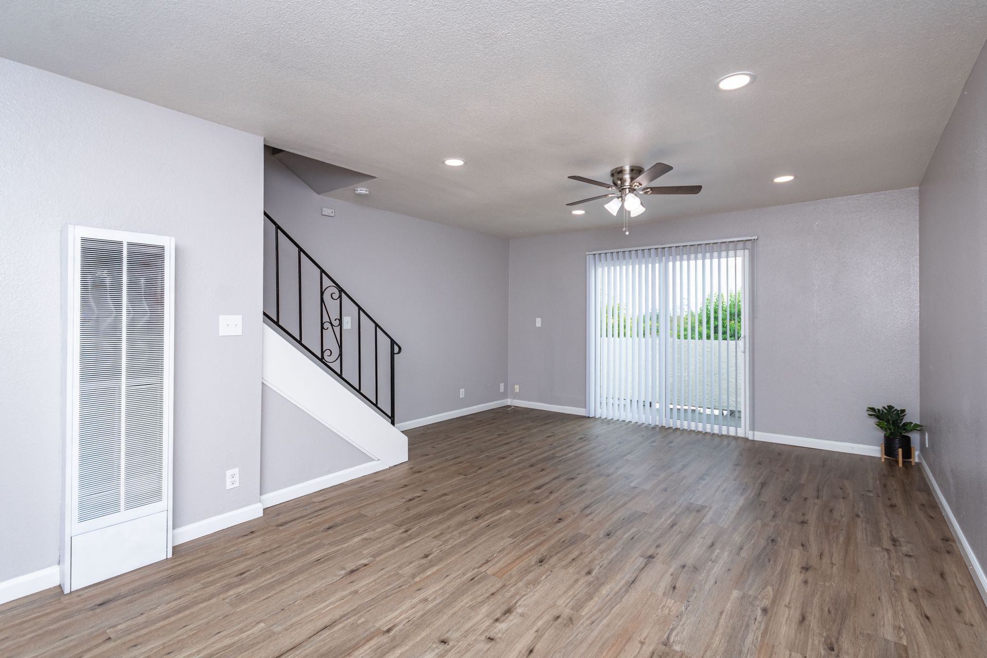 Empty gray living room with wood floors, staircase, ceiling fan, and sliding glass door.