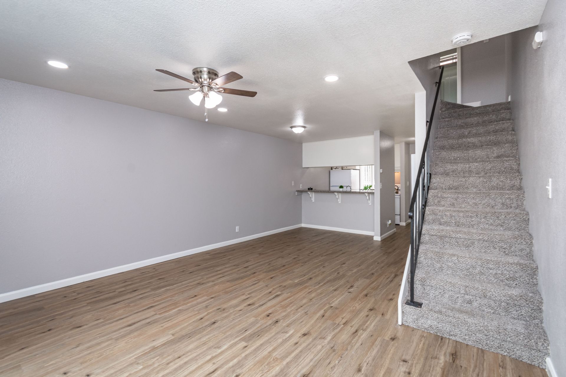 Empty living room with gray walls, wood-look floor, ceiling fan, kitchen pass-through, and carpeted stairs.