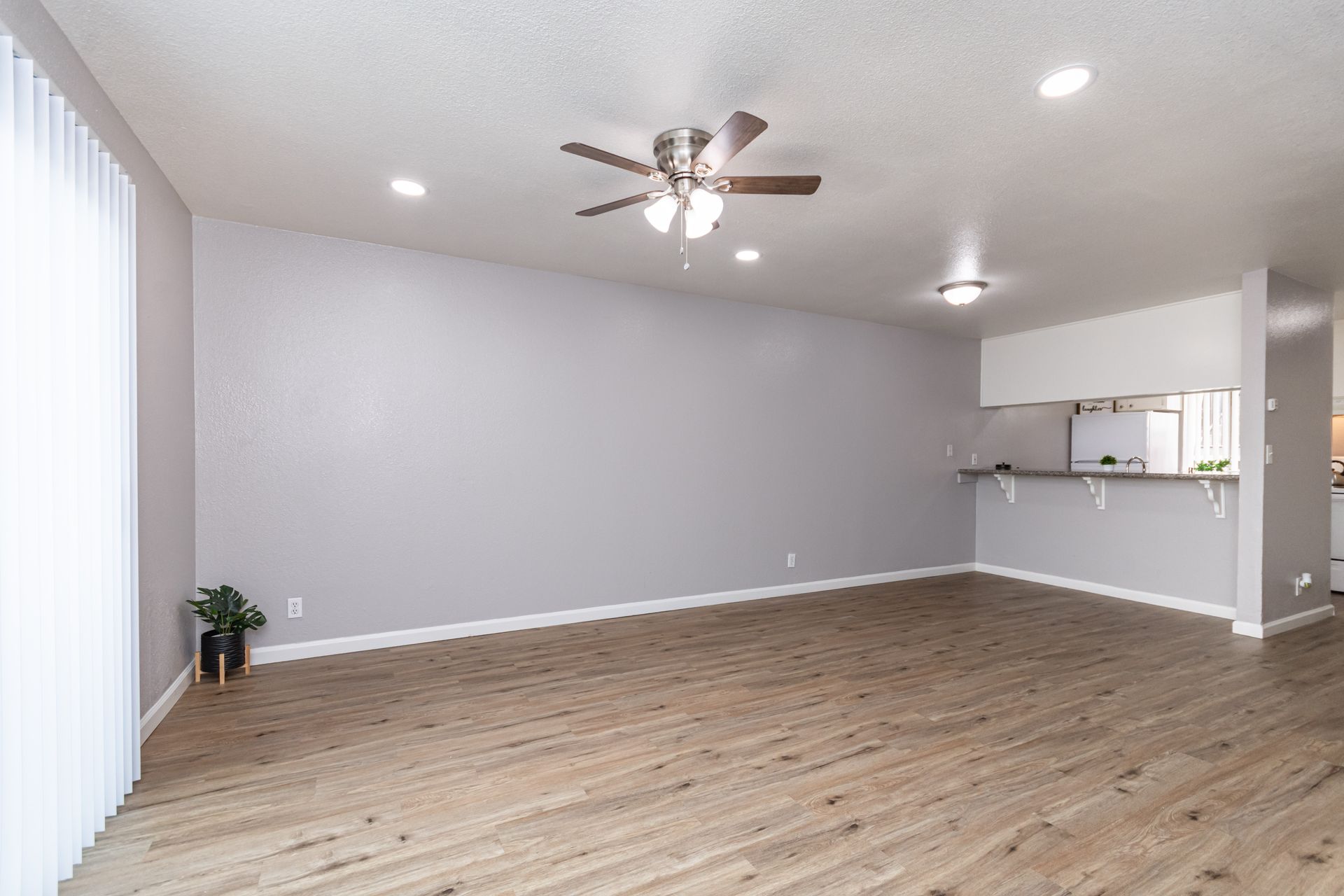 Spacious empty living room with gray walls, wood flooring, ceiling fan, and open kitchen area.