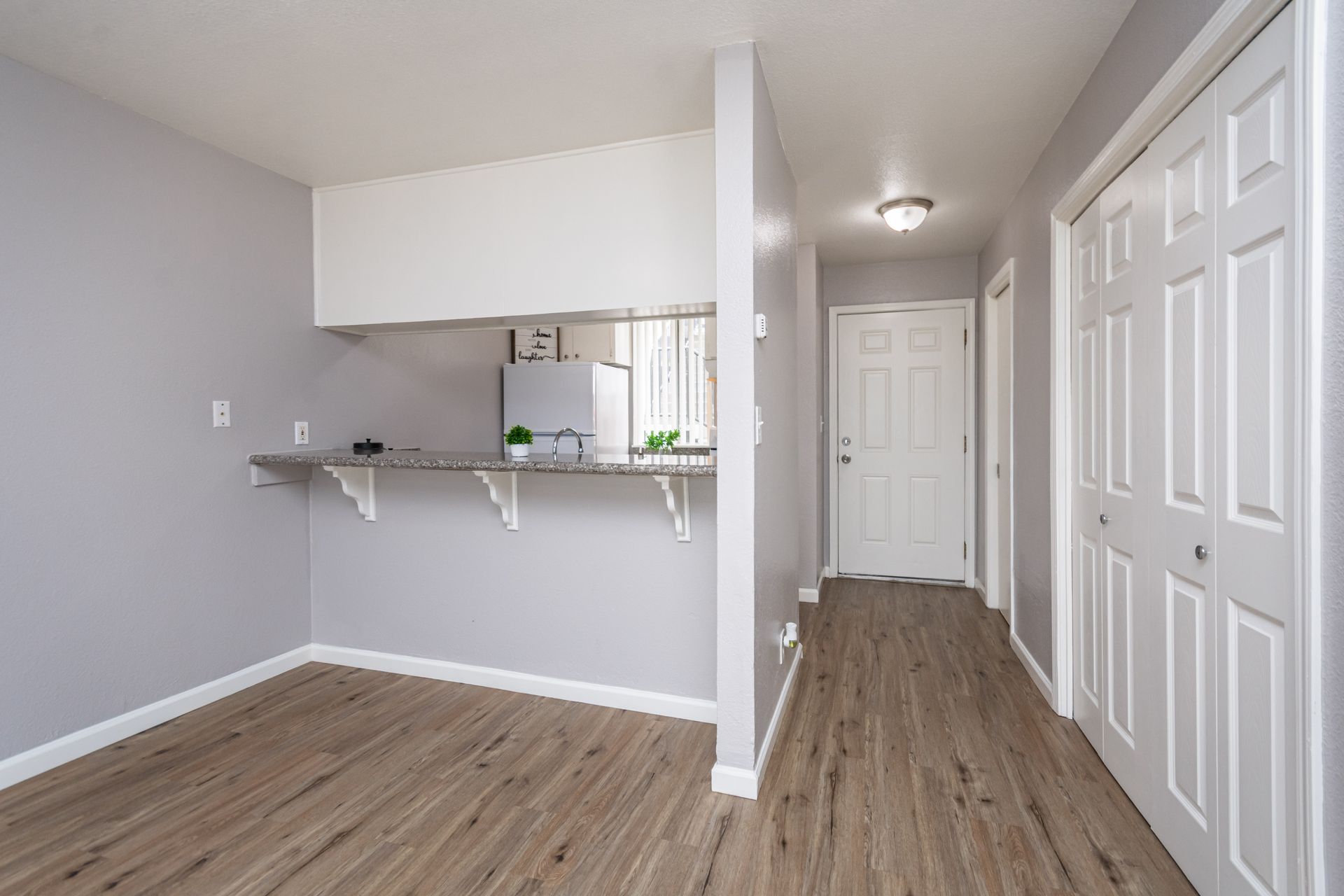 Empty apartment hallway with light wood floors, gray walls, and white doors and kitchen pass-through.