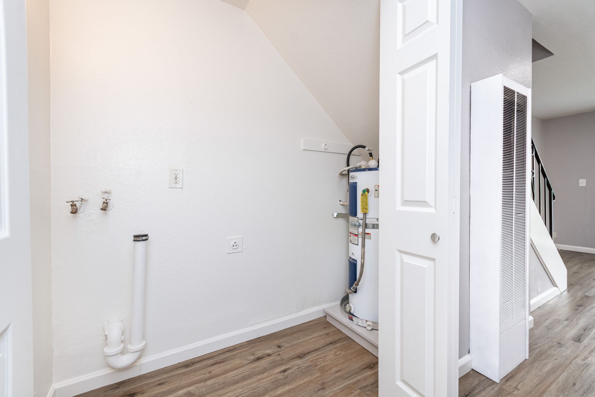 Empty laundry room with white walls, wood floor, sink plumbing, and a utility unit behind an open door