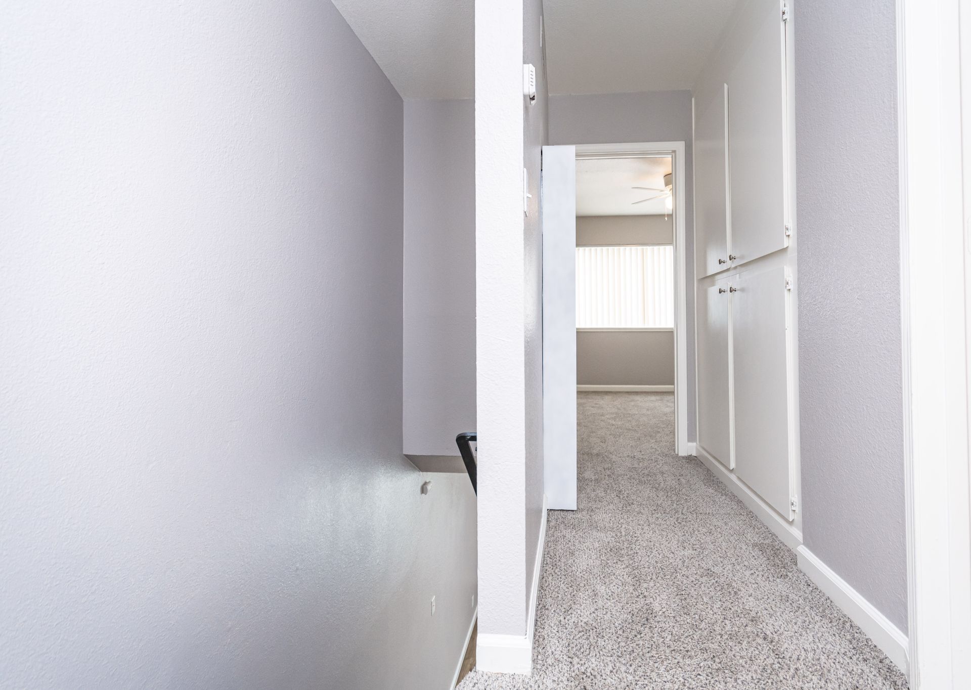 Narrow carpeted hallway with white walls, an open door, and a bright room at the end.