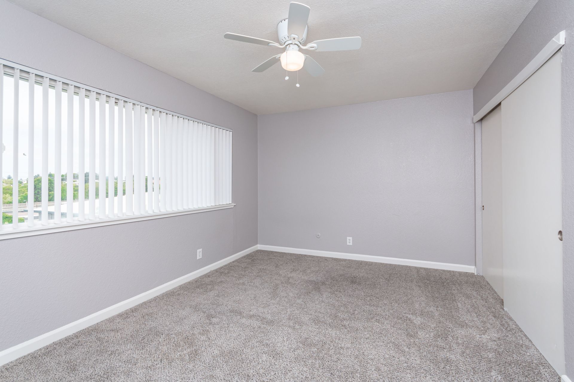 Empty gray-carpeted bedroom with white walls, a window with blinds, and a ceiling fan lights fixture