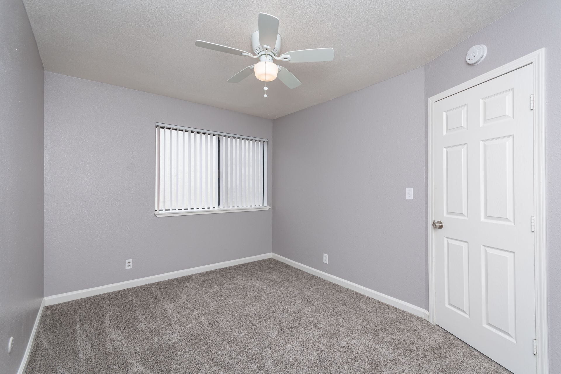 Empty light gray bedroom with white ceiling fan, carpet, window blinds, and closet door