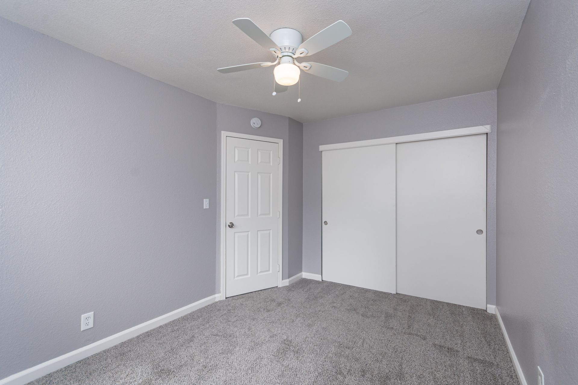Empty gray bedroom with carpet, white ceiling fan, closet doors, and a single window.