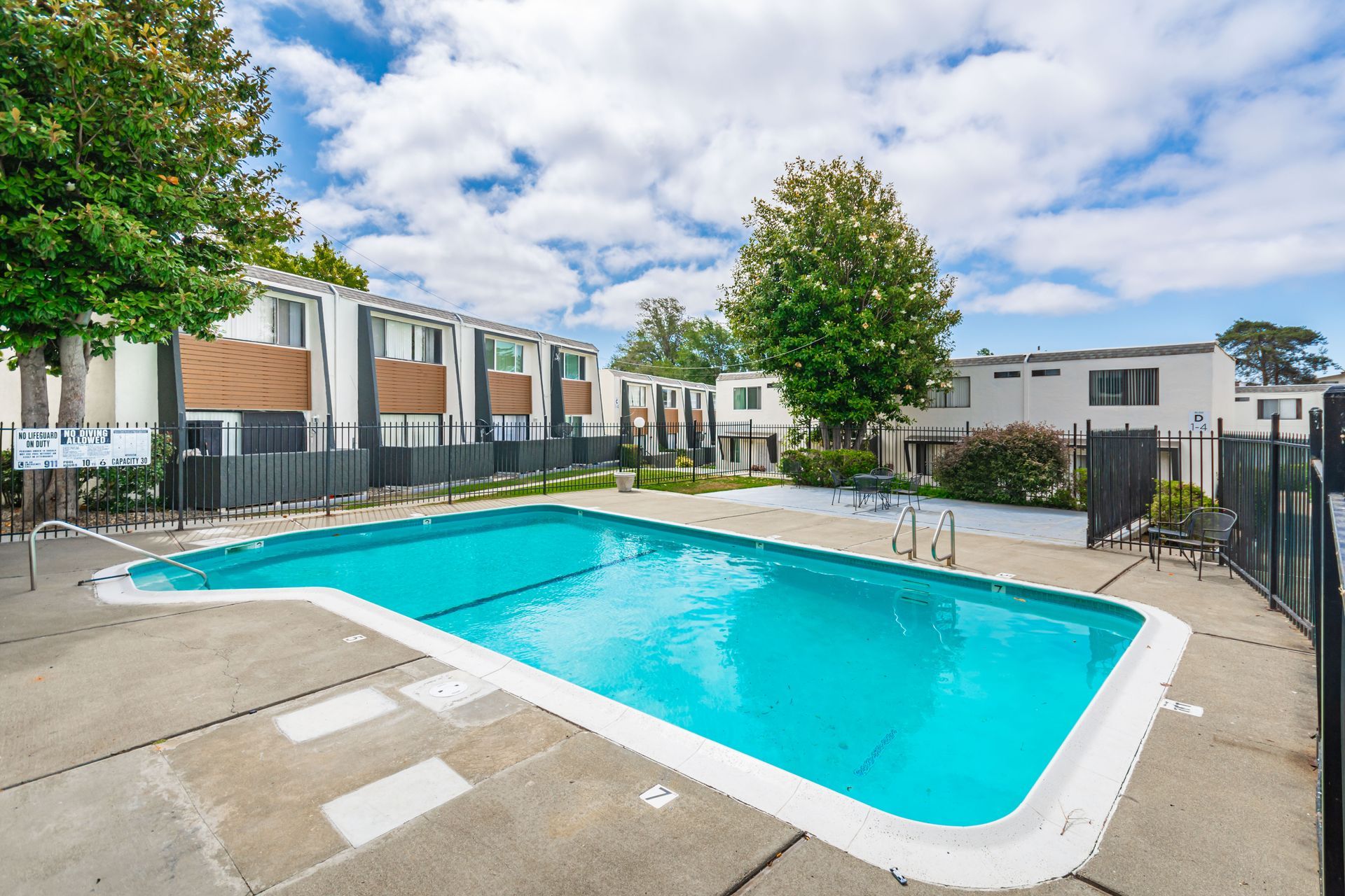 Outdoor apartment pool with turquoise water, white deck, black fence, and townhomes under a cloudy sky