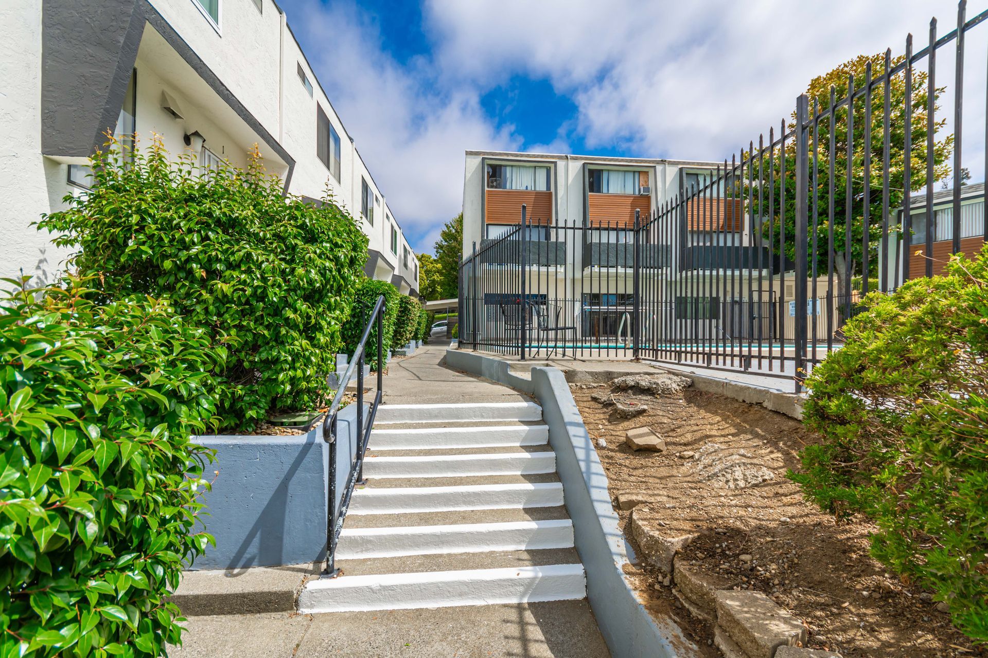 Concrete stairs between apartment buildings, bordered by greenery and a black fence under a blue sky.