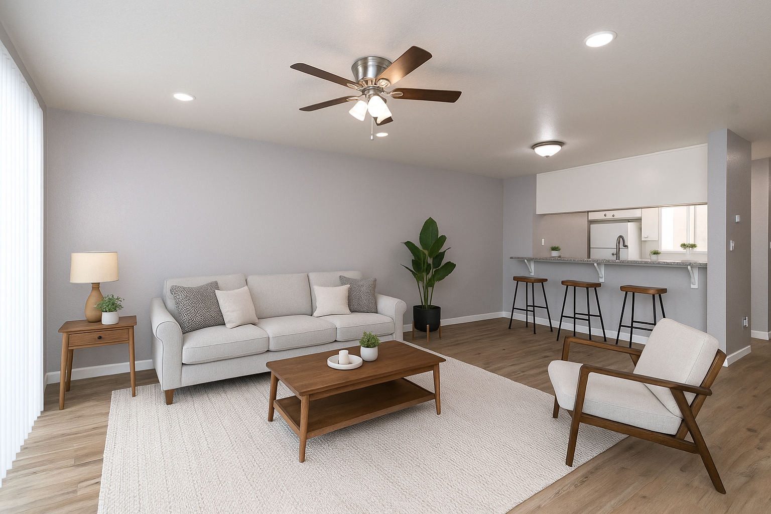 Bright living room with gray sofa, wooden coffee table, armchair, ceiling fan, and open kitchen bar stools.