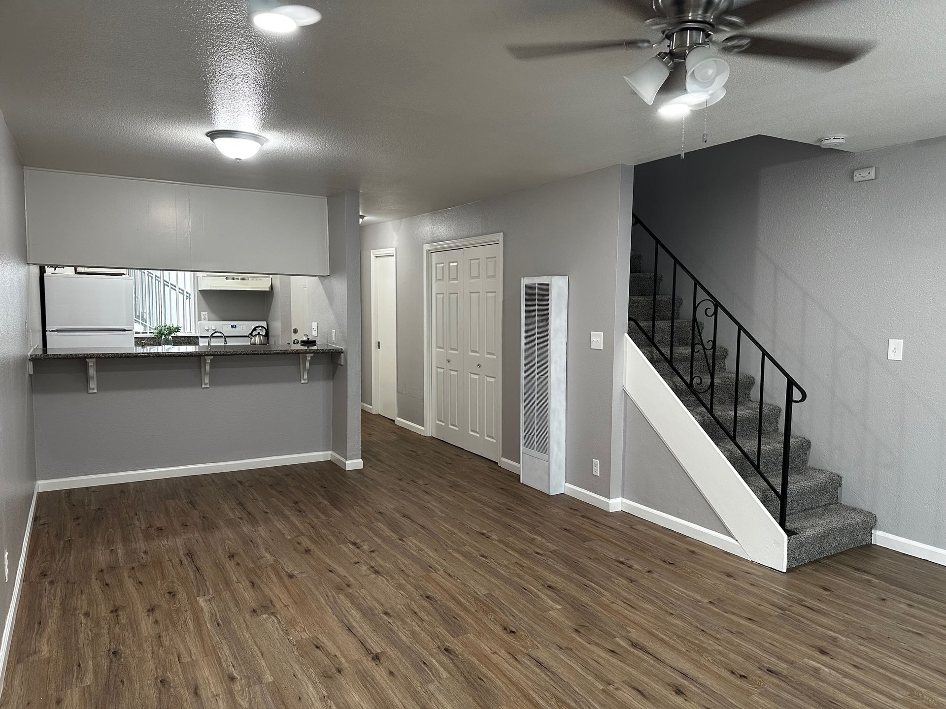 Empty living room with wood floors, ceiling fans, open kitchen pass-through, and carpeted stairs.