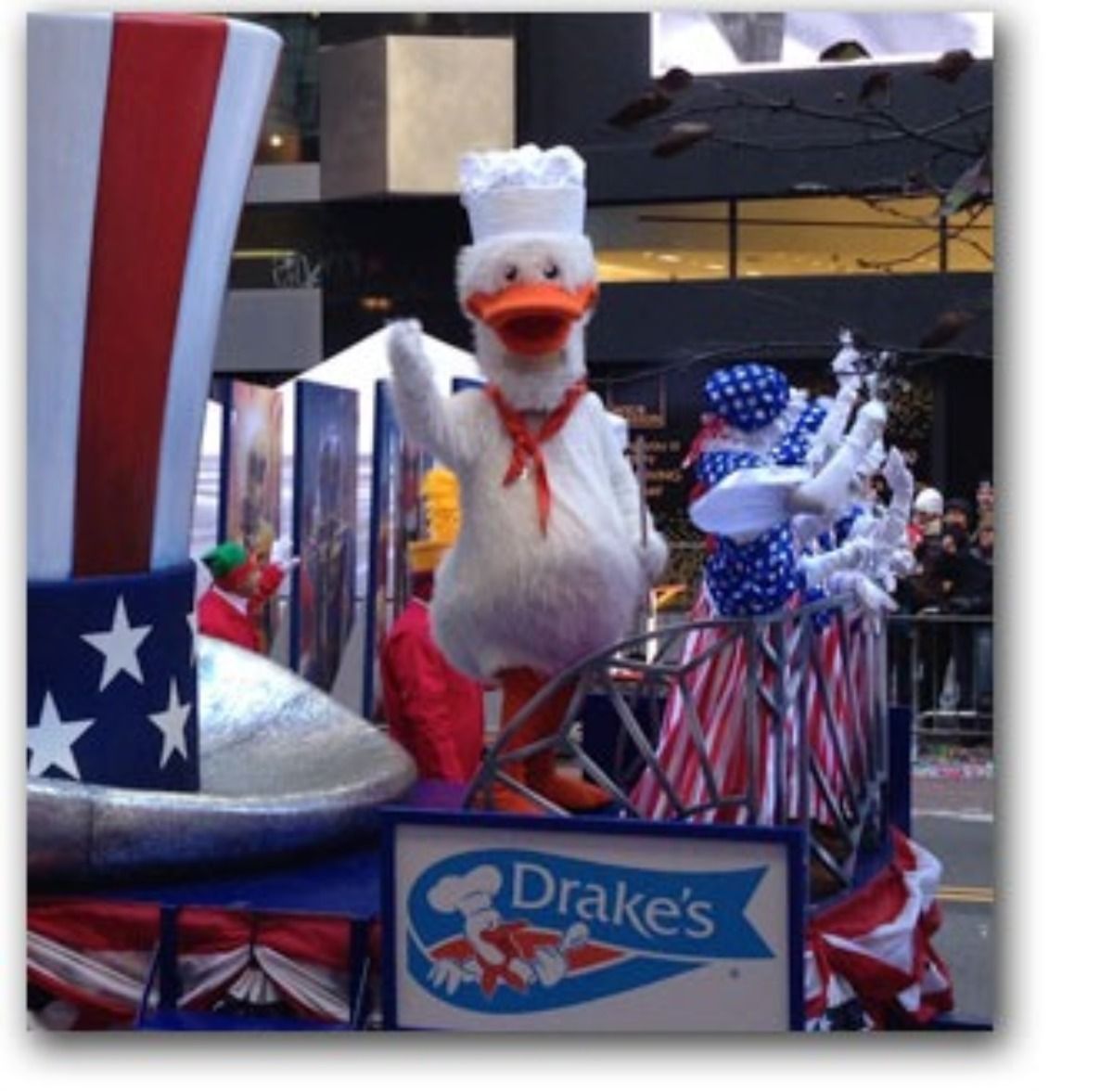 A costumed duck mascot in a chef’s hat waves from a patriotic Drake's parade float decorated with stars and stripes.