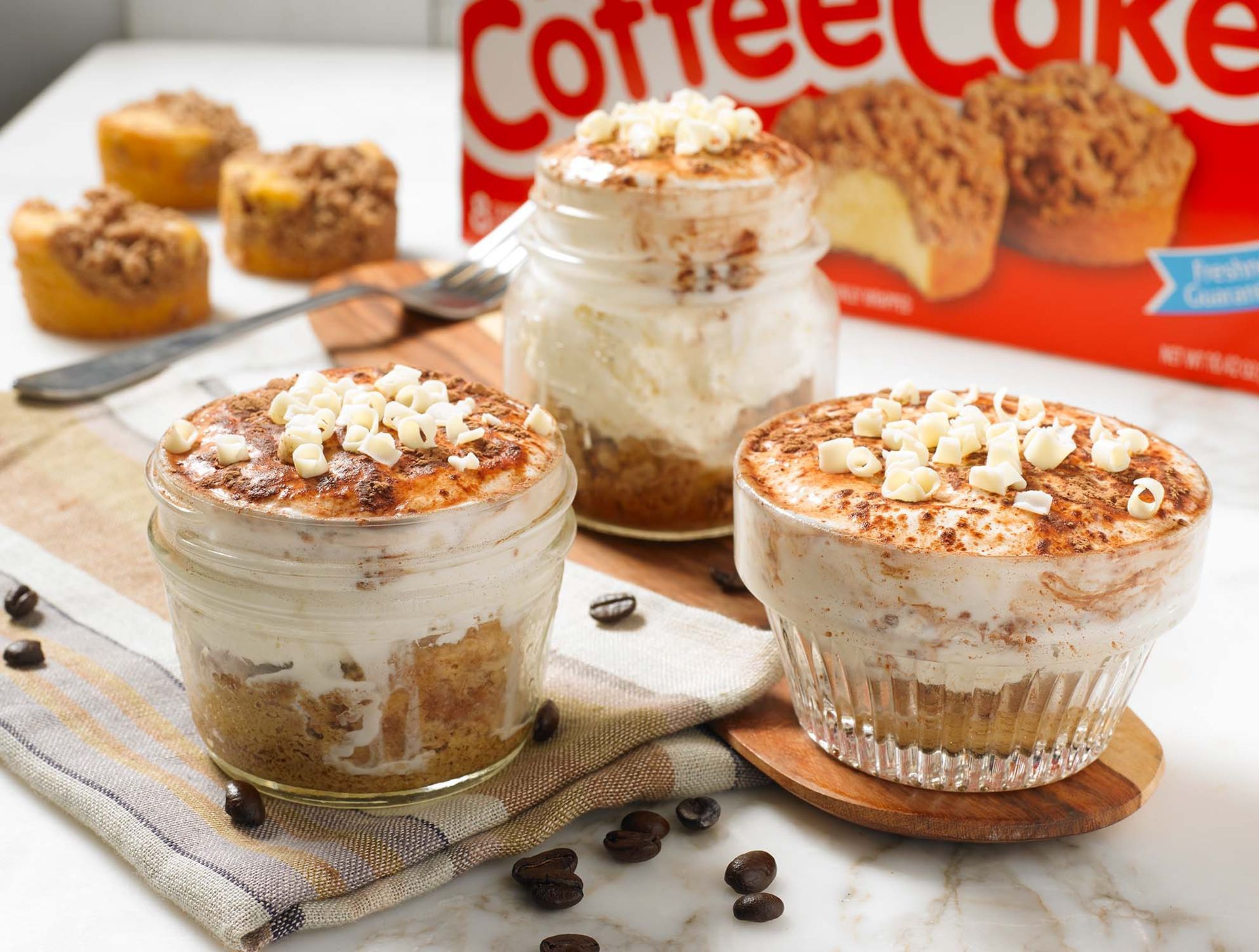 Three glass jars filled with layered coffee cake and cream desserts, garnished with white chocolate chips on a table.