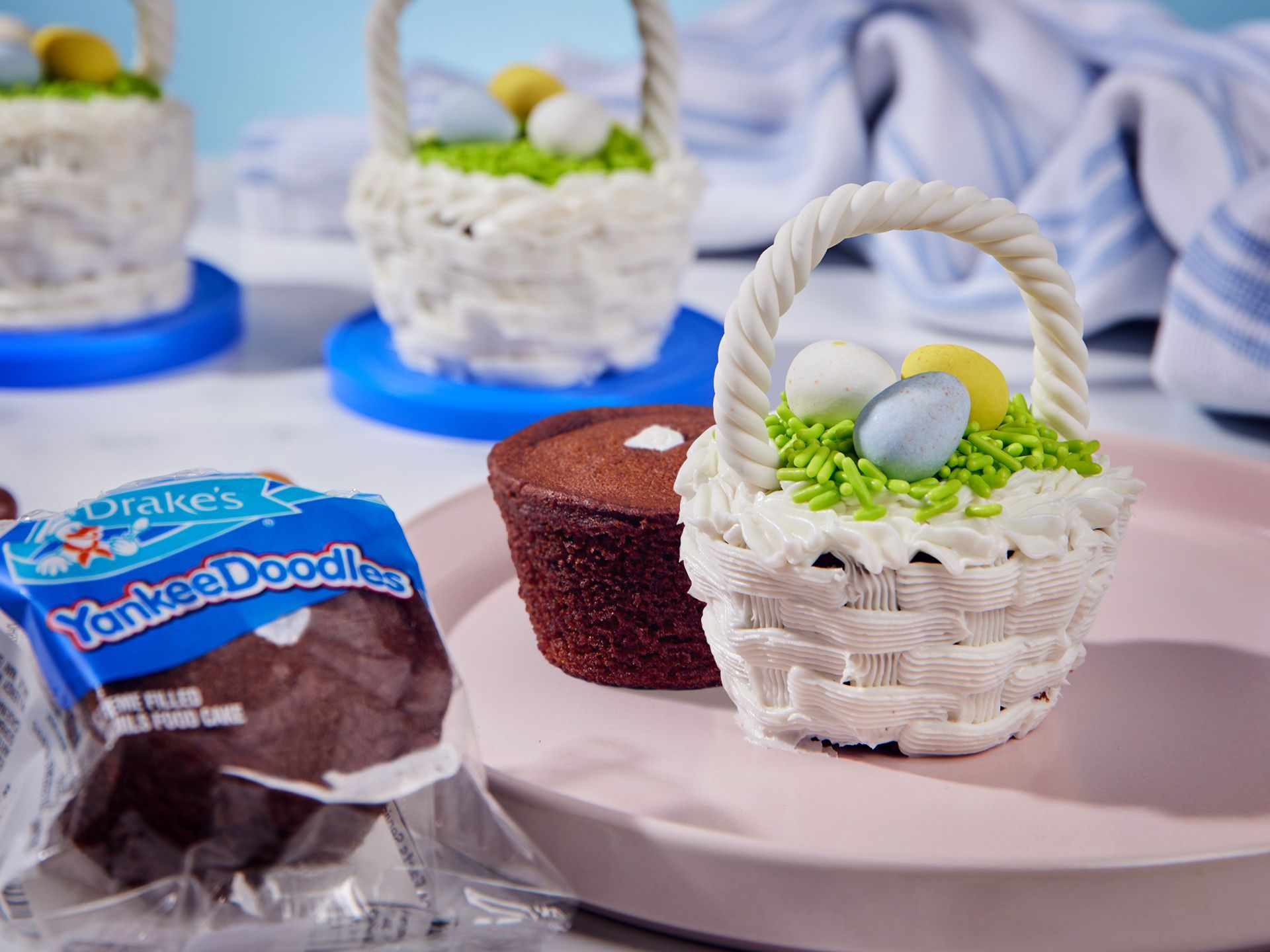 Cupcakes decorated as Easter baskets with white frosting and candy eggs, next to a package of Drake’s YankeeDoodles cakes.