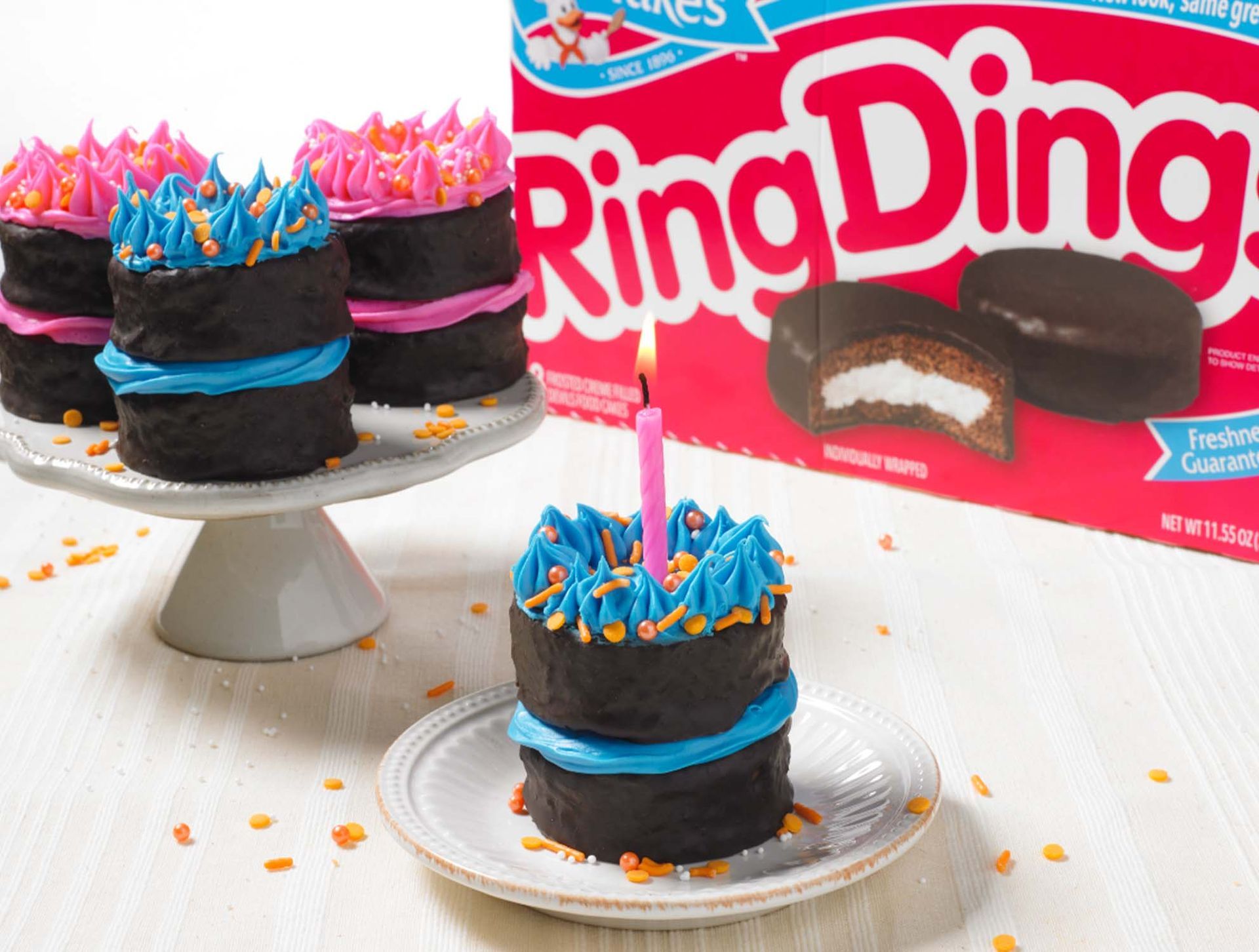 Three mini chocolate cakes decorated with pink or blue icing sit on white plates, with a Ring Ding box in the background.