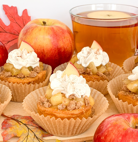 Apple pie cupcakes topped with whipped cream and crumbles, arranged near fresh apples, autumn leaves, and apple cider.