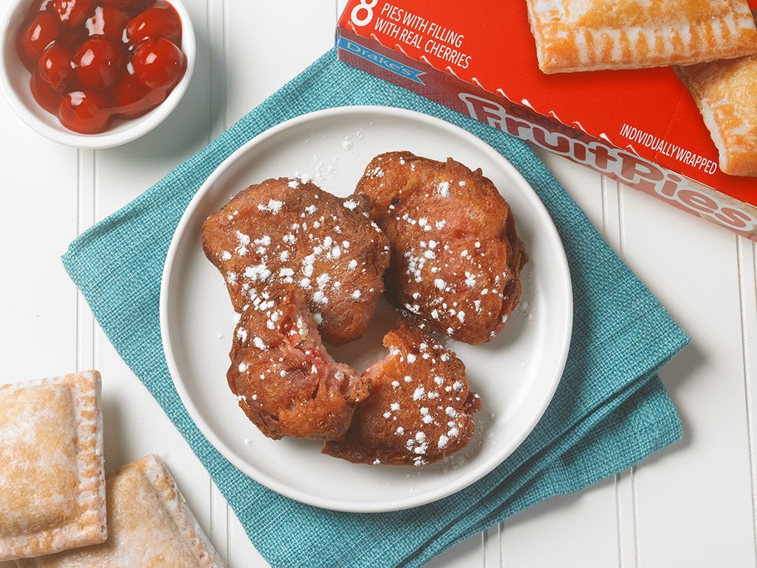 A plate of four powdered sugar-topped fried pastries on a blue napkin, with a bowl of cherries and boxed fruit pies nearby.