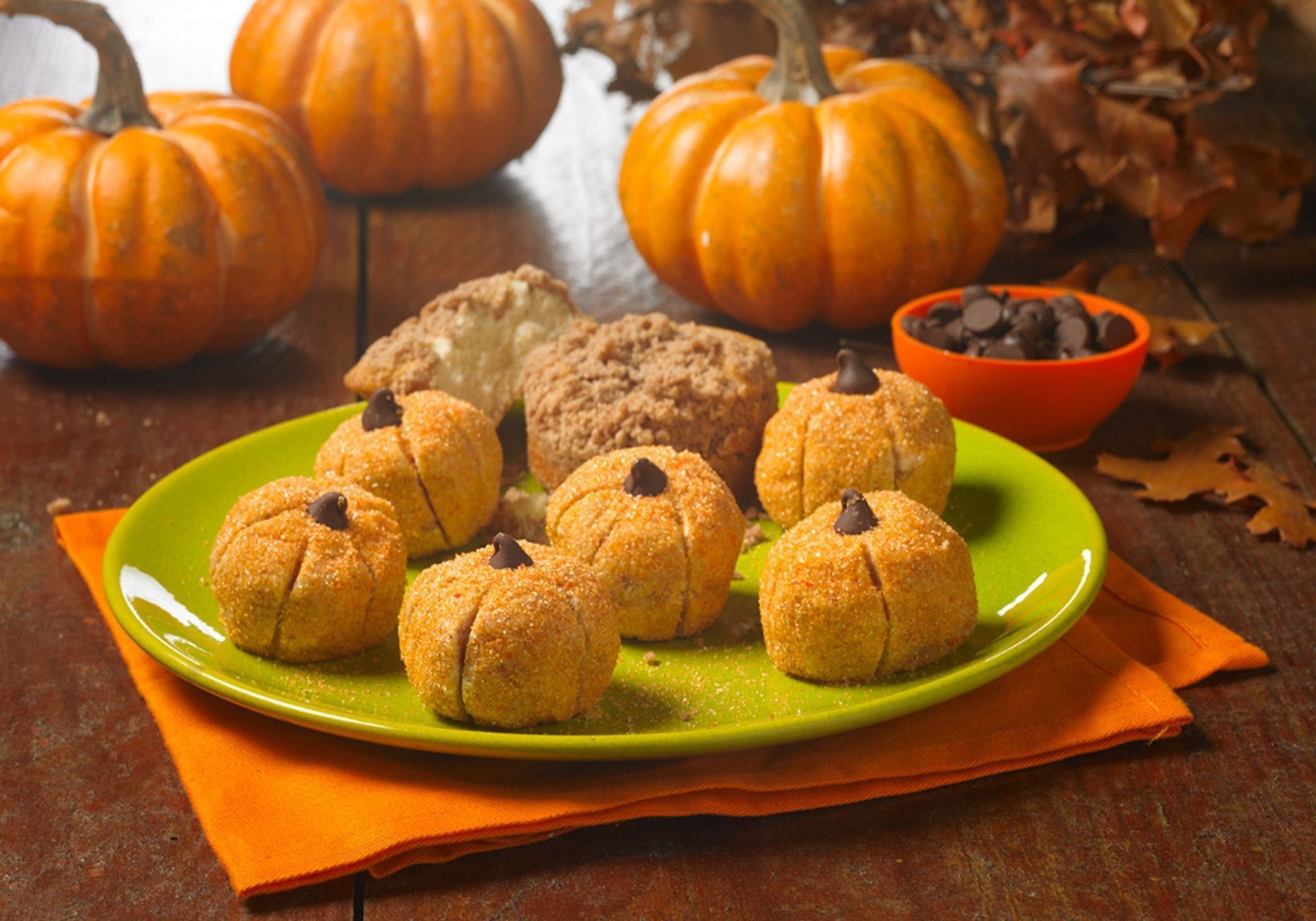 A lime green plate holding pumpkin-shaped cookies with chocolate chip stems, set on a wooden table with fall pumpkins.