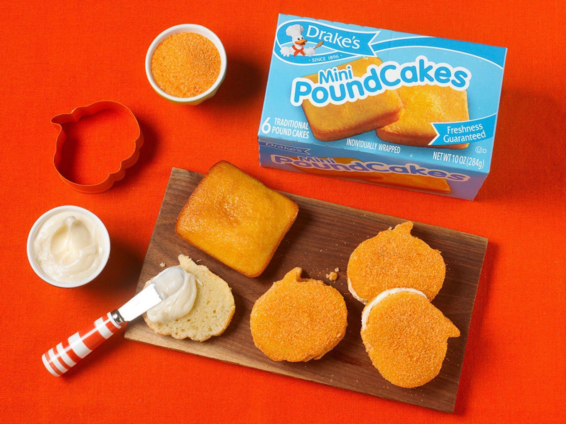A box of Drake’s Mini Pound Cakes, orange sprinkles, a pumpkin cookie cutter, and frosted cake slices on a wooden board.