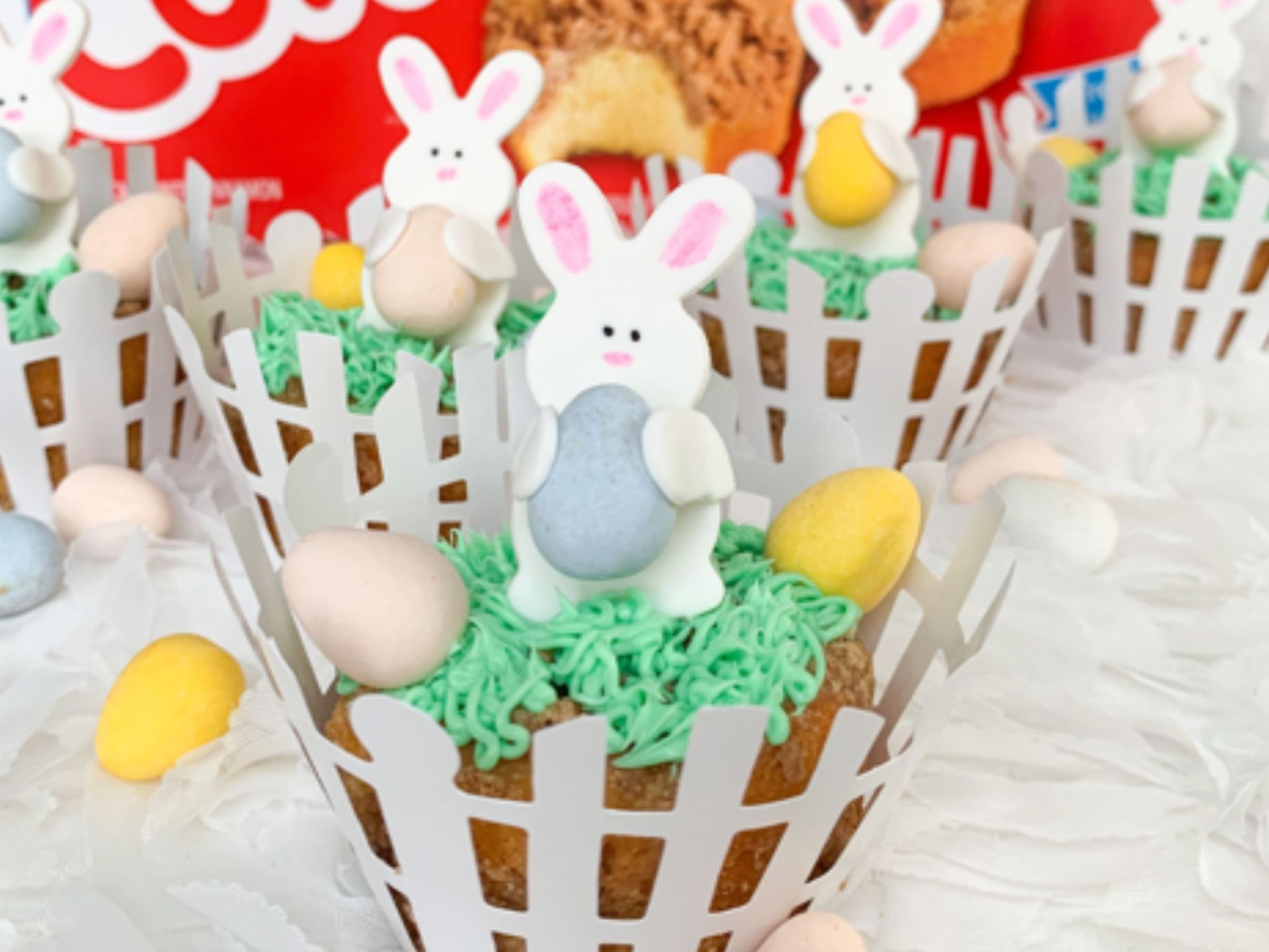 Easter cupcakes in white picket fence wrappers, topped with green frosting, candy eggs, and fondant bunnies.