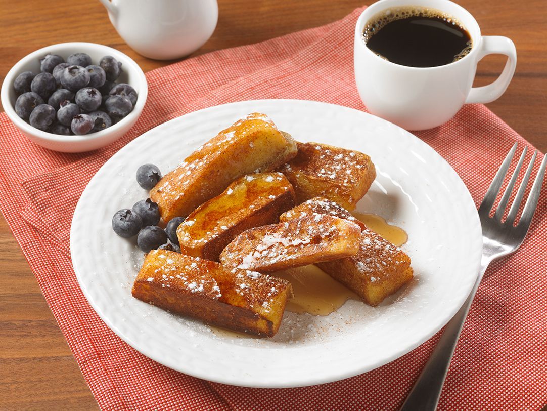 A plate of French toast sticks sprinkled with powdered sugar and syrup, with a side of blueberries and a cup of coffee.