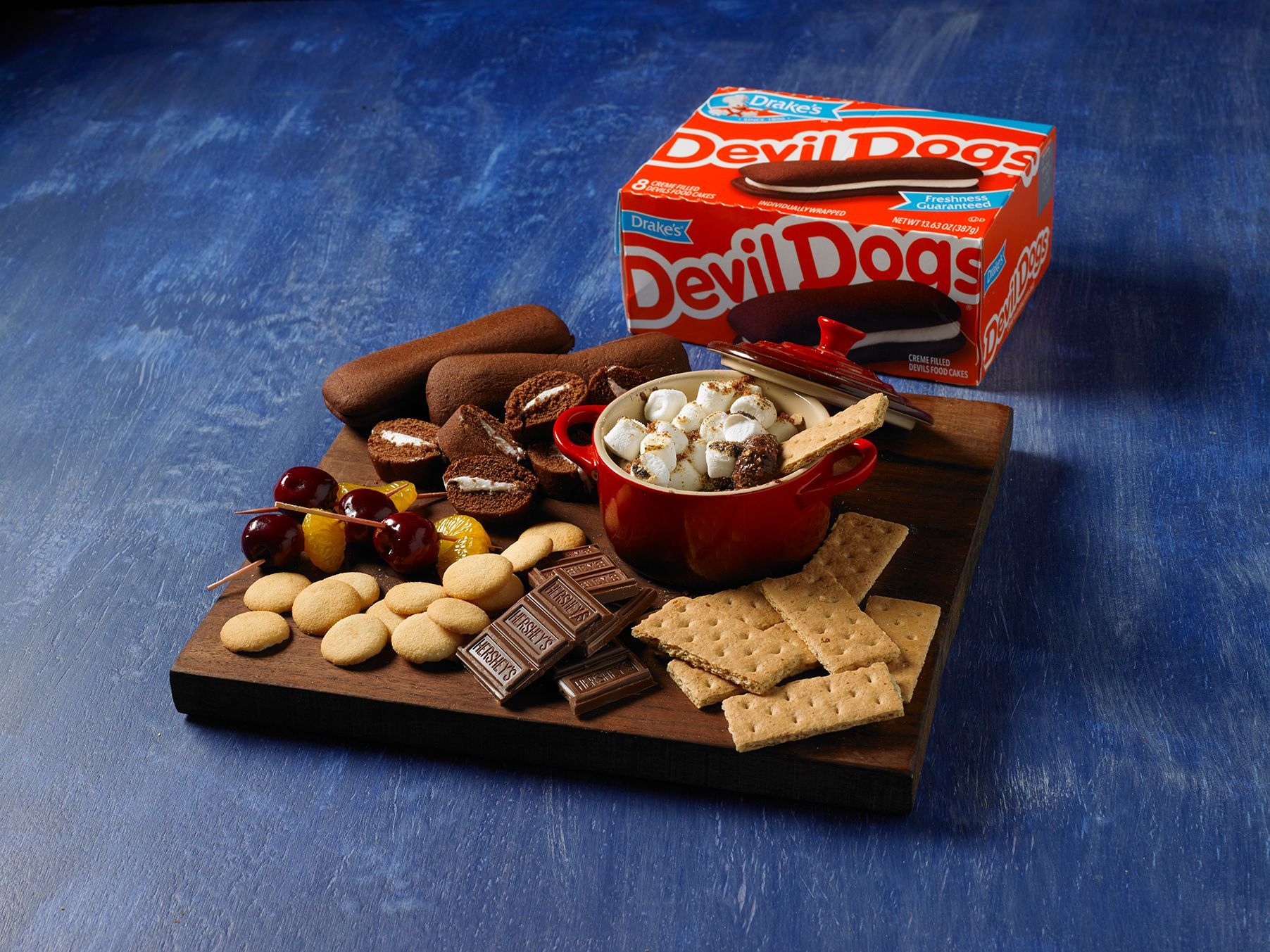 A wooden board with Devil Dogs, s'mores dip, graham crackers, cookies, cherries, and chocolate against a blue background.