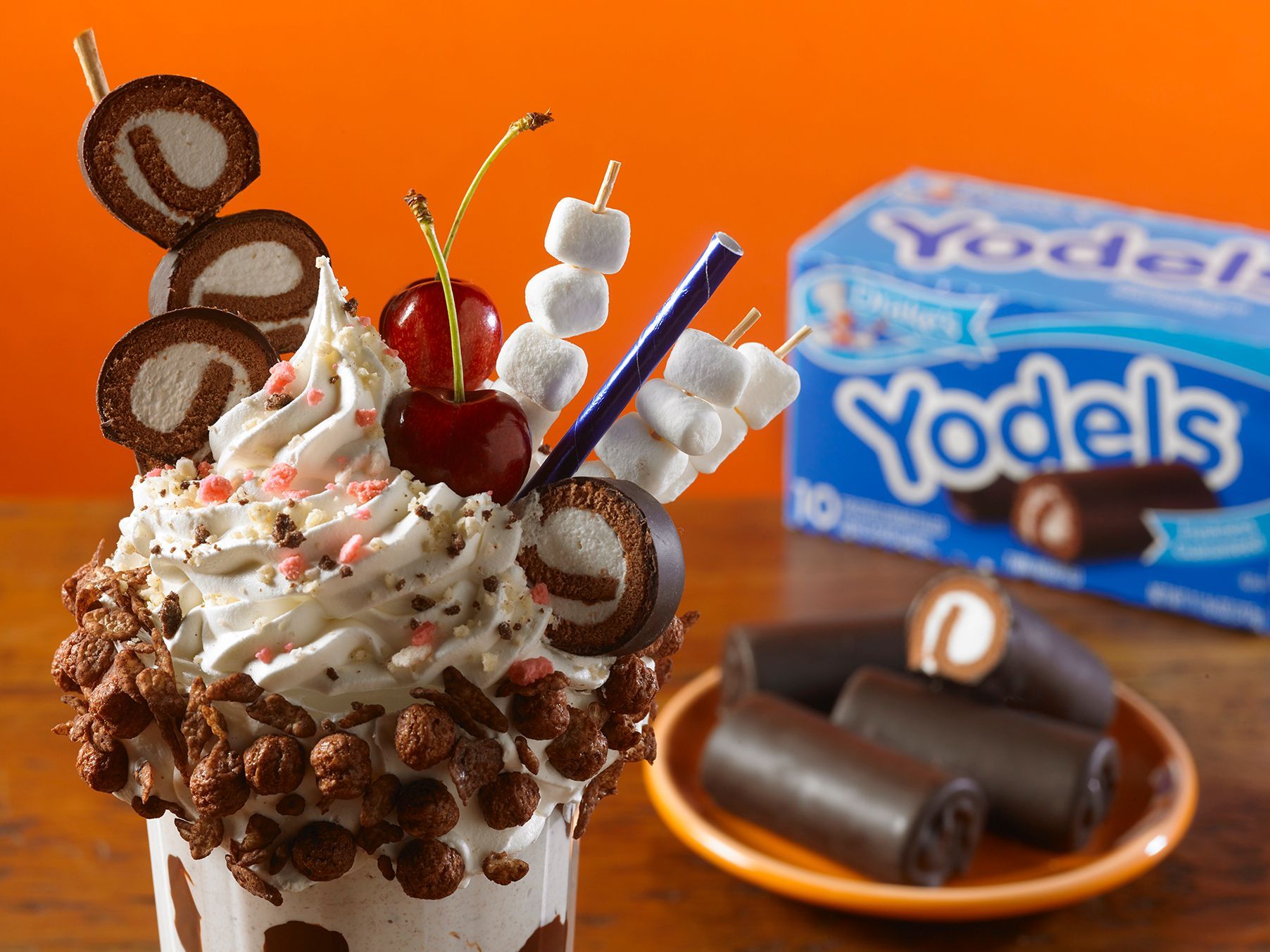 A decadent milkshake topped with Yodels, whipped cream, cherries, and marshmallows, with a box of Yodels in the background.