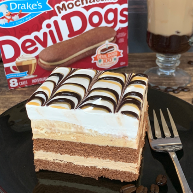 A layered chocolate and mocha cake slice on a black plate, next to a box of Drake’s Mocha Creme Devil Dogs and a drink.