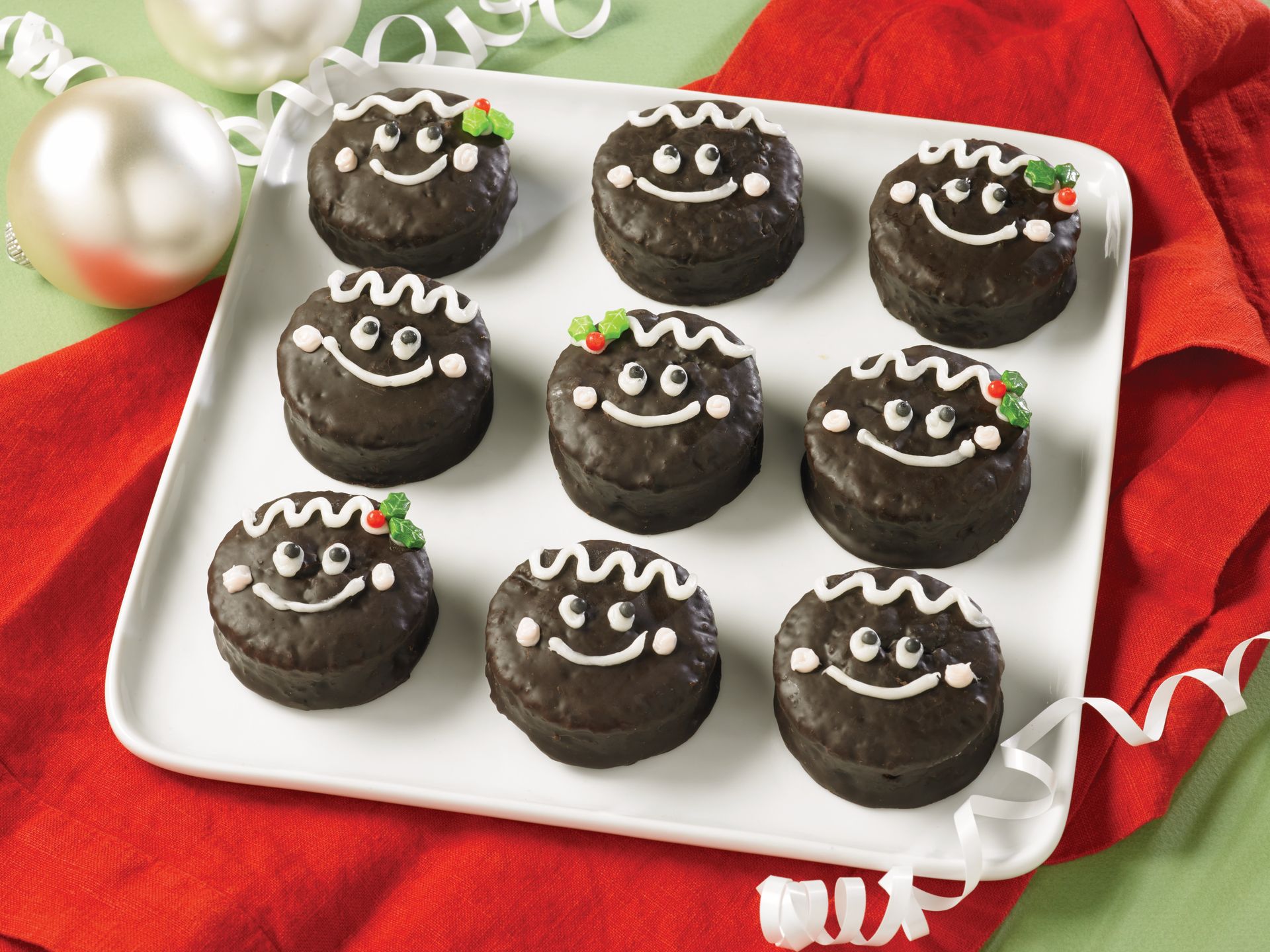 Nine round, chocolate-covered treats decorated with white icing faces and small holly sprigs, arranged on a white plate.