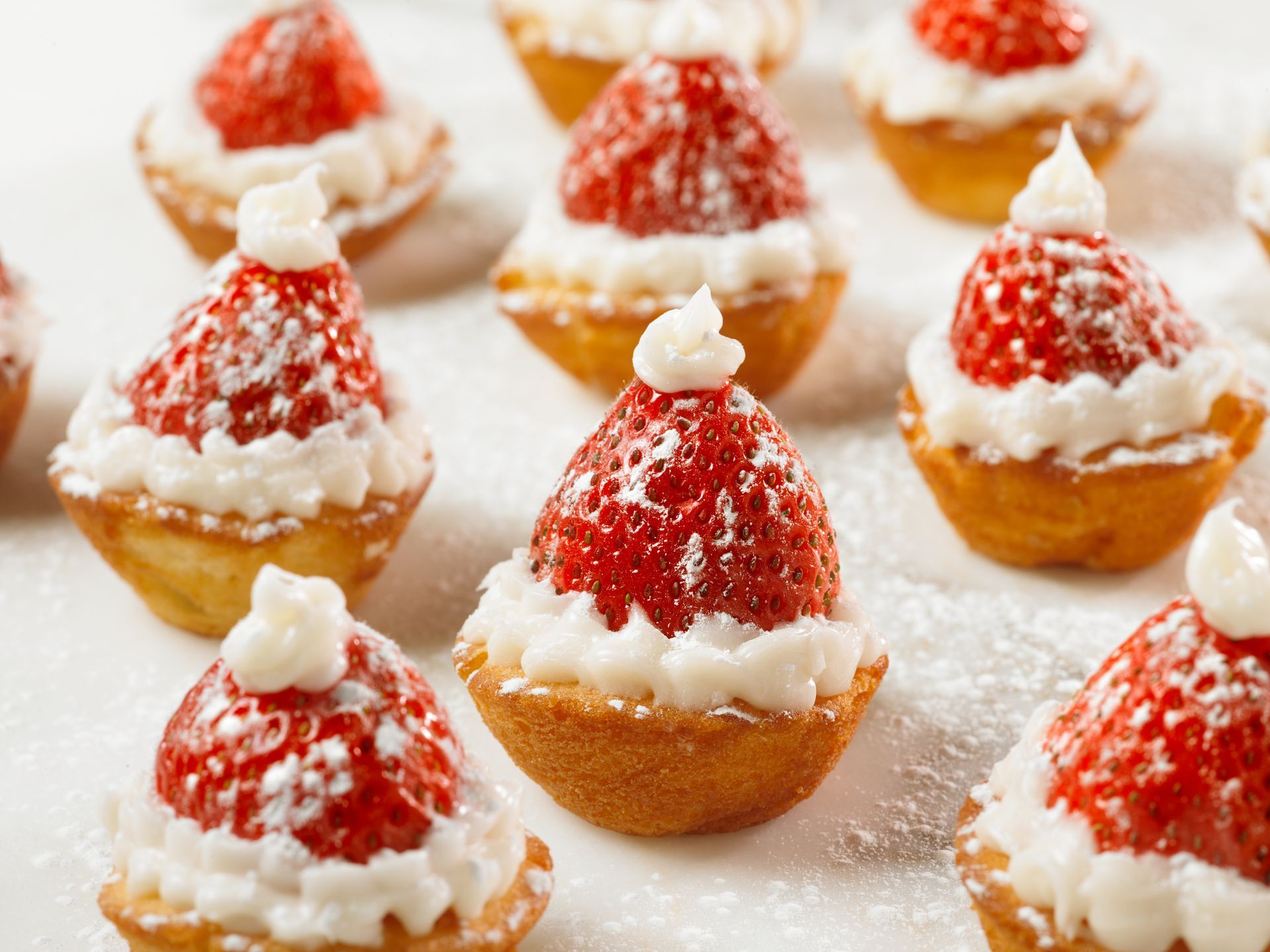 A collection of strawberry Santa hat mini tarts with whipped cream, dusted with powdered sugar on a white surface.