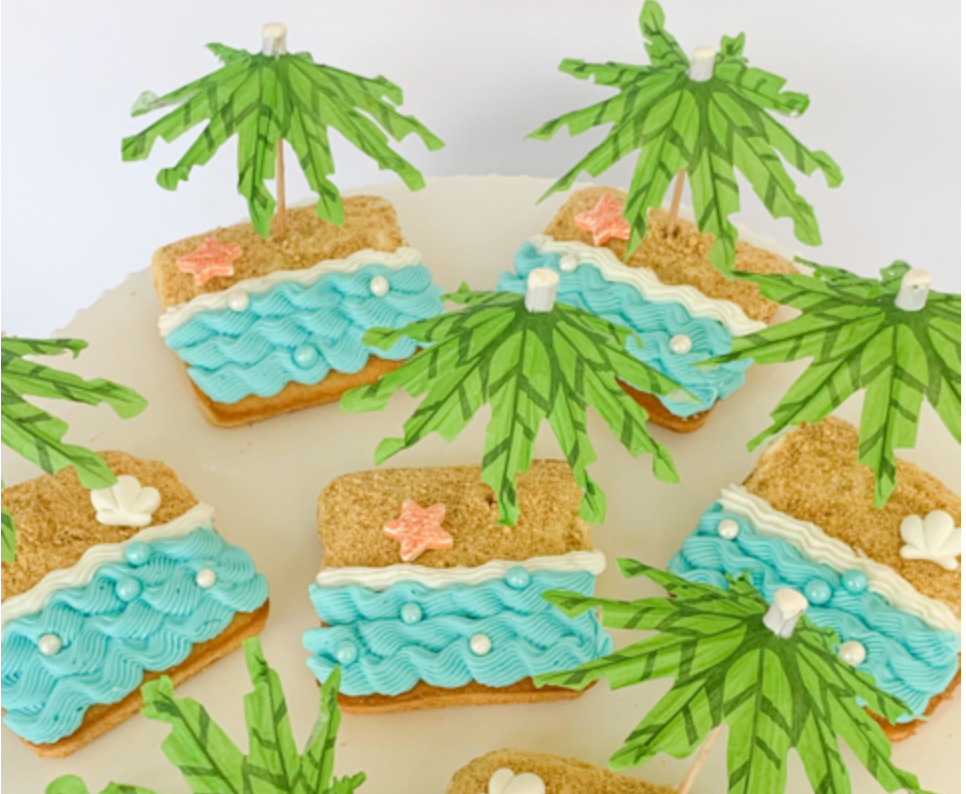 Square cookies decorated as beaches with blue frosting waves, sandy graham cracker crumbs, and miniature paper palm trees.