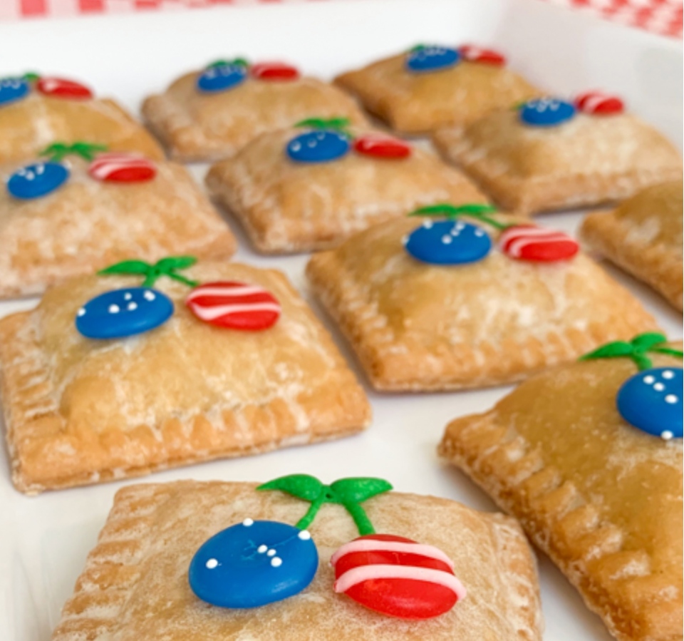 Square pastries decorated with blue and red candy cherries and green frosting stems on a white tray.
