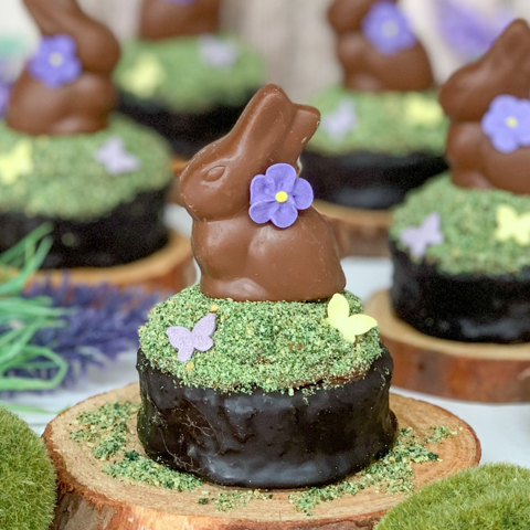 Chocolate Easter treats with bunny figurines, purple flowers, and butterfly sprinkles on green coconut grass, on wood bases.